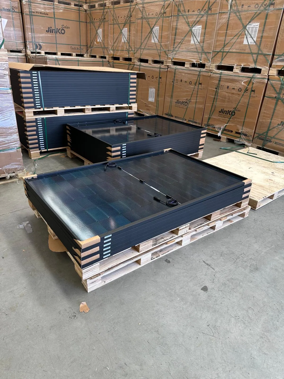 Stacks of black-framed solar panels on timber pallets in front of boxed Jinko Solar modules